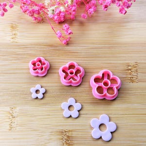 Peut inclure: Plusieurs emporte-pièces en forme de fleurs roses et blanches de différentes tailles sont disposés sur une surface en bois. Les emporte-pièces sont en plastique et présentent un design simple à cinq pétales. Des fleurs roses et de la verdure sont en arrière-plan.