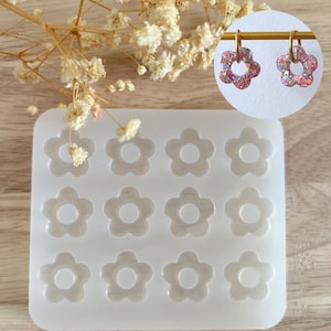 Peut inclure: Un moule en silicone transparent avec douze cavités en forme de fleur. Deux boucles d'oreilles en forme de fleur avec des paillettes sont affichées à droite. Les boucles d'oreilles ont un cerceau doré et sont remplies de paillettes roses, violettes et argentées. Des fleurs séchées sont en arrière-plan.