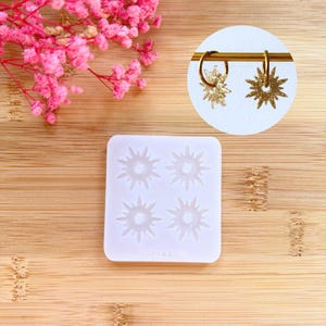 Op de afbeelding: Een witte siliconen mal met vier sterrenvormen, naast een paar goudkleurige sterrenoorbellen. Roze bloemen bevinden zich in de linkerbovenhoek. De oorbellen hangen aan een gouden staaf.