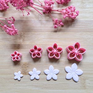 Puede incluir: Colección de cortadores de galletas con forma de flor rosa y blanca de varios tamaños. Los cortadores están dispuestos sobre una superficie de madera marrón claro, con flores rosas en el fondo.