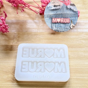 Peut inclure: Un moule en silicone blanc avec le mot "MOM" et une forme de cœur, avec le texte "TYLIA&CO" en bas. Un patch en jean avec le mot "MORUE" est également montré. Des fleurs roses sont en arrière-plan.