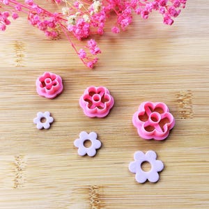 Peut inclure: Des découpoirs à biscuits et des fleurs découpées en forme de fleurs roses et lavande sont disposés sur une surface en bois. Les découpoirs, de différentes tailles, ont un trou central. Des fleurs roses séchées sont en arrière-plan.