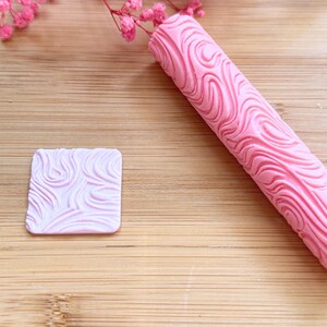 Peut inclure: Un rouleau à pâtisserie rose texturé et un petit tampon carré, tous deux avec un motif tourbillonnant. Ces outils, probablement pour la pâtisserie ou l'artisanat, sont en matériau lisse et solide. Ils sont présentés sur une surface en bois.