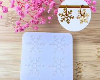 Op de afbeelding: Een doorzichtige siliconen mal met vier sneeuwvlokontwerpen en een paar gouden sneeuwvlok oorbellen. Roze bloemen op de achtergrond. De mal heeft de tekst "TYLIA&CO" onderaan.