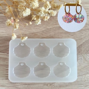 Peut inclure: Un moule en silicone transparent avec six cavités en forme de coquillage, chacune avec un petit trou en haut. Une paire de boucles d'oreilles coquillage scintillantes finies avec des cerceaux dorés est montrée en haut à droite. Fleurs séchées en arrière-plan.