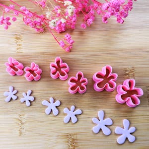 Könnte beinhalten: Eine Sammlung von rosa und lavendelfarbenen Ausstechern und Fondant-Ausstechern in Blumenform. Die Ausstecher gibt es in verschiedenen Größen und Designs, ideal zum Backen und Basteln. Rosa Trockenblumen sind im Hintergrund, alles auf einer Holzoberfläche.