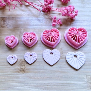 Puede incluir: Un conjunto de cortadores de galletas rosas en forma de corazón, de varios tamaños, con un diseño acanalado. Los cortadores están dispuestos sobre una superficie de madera, con acentos florales rosas. Ideal para hornear y manualidades.