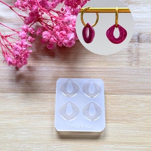 Puede incluir: Pendientes de aro dorados con colgantes ovalados de color rosa oscuro. También se ve un molde de silicona transparente con cuatro cavidades en forma de pendientes. Flores rosas en el fondo, todo sobre una superficie de madera clara.