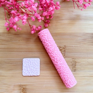 Peut inclure: Un rouleau à pâtisserie rose texturé et un petit tampon carré avec un motif similaire. Les articles sont probablement destinés à la pâtisserie ou à l'artisanat, avec un motif léopard. Des fleurs roses sont en arrière-plan.
