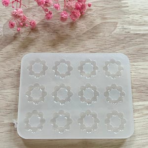 Peut inclure: Un moule en silicone transparent et rectangulaire avec douze cavités en forme de fleur. Le moule est conçu pour la fabrication de petits éléments floraux décoratifs. Des fleurs roses sont en arrière-plan.