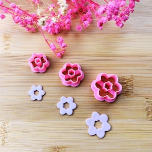 Peut inclure: Ensemble de découpoirs à biscuits en forme de fleurs roses et violet clair de différentes tailles. Les découpoirs sont en plastique et disposés sur une surface en bois, avec des fleurs roses en arrière-plan.