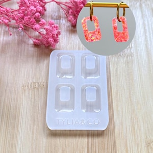 Peut inclure: Boucles d'oreilles rectangulaires roses et oranges à paillettes, montées sur des anneaux dorés. Un moule en silicone transparent avec quatre cavités rectangulaires est au premier plan. Le moule porte le texte "TYLIA&CO".