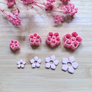 Puede incluir: Cortadores de galletas rosas con forma de flor en varios tamaños, dispuestos sobre una superficie de madera. Los cortadores son de plástico y tienen un diseño floral detallado. Flores rosas secas en el fondo.