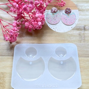 May include: A clear silicone earring mold with two earring shapes, and a pair of finished glitter earrings. The earrings are pink and white with a glittery finish. Pink dried flowers are in the background. The mold has the text "TYLIA&CO".