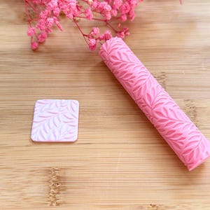 Puede incluir: Un rodillo rosa con estampado de hojas y un pequeño sello cuadrado están sobre una superficie de madera. El rodillo es cilíndrico y el sello es cuadrado. Hay flores rosas en el fondo. Las herramientas son probablemente para hornear o manualidades.