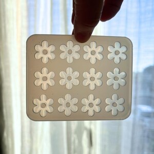 Puede incluir: Un molde de silicona transparente y rectangular con 12 cavidades en forma de flor. El molde est&aacute; sostenido por una mano y est&aacute; dise&ntilde;ado para la elaboraci&oacute;n o creaci&oacute;n de peque&ntilde;os art&iacute;culos con tem&aacute;tica floral. Las cavidades est&aacute;n espaciadas y detalladas.