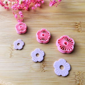 May include: A collection of pink and lavender flower-shaped cookie cutters and fondant molds. The cutters are made of pink plastic, while the fondant molds are a soft lavender color. The cutters and molds are arranged on a wooden surface, with pink flowers in the background.