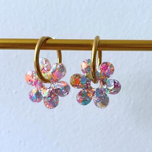Puede incluir: Un par de pendientes con forma de flor con aros dorados. Los p&eacute;talos de la flor est&aacute;n rellenos de purpurina de colores, incluyendo rosa, naranja, azul y morado. Los pendientes cuelgan de una barra dorada sobre un fondo blanco.