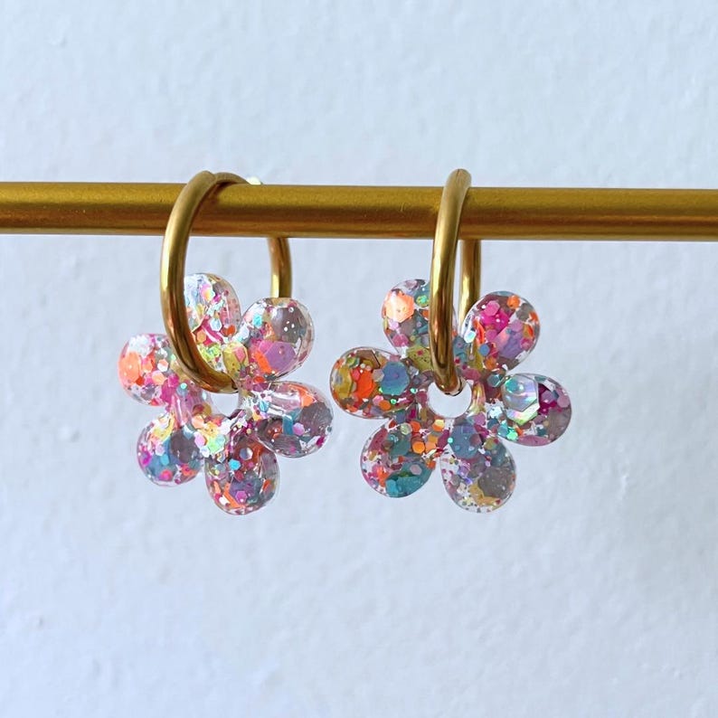 Puede incluir: Un par de pendientes con forma de flor con aros dorados. Los p&eacute;talos de la flor est&aacute;n rellenos de purpurina de colores, incluyendo rosa, naranja, azul y morado. Los pendientes cuelgan de una barra dorada sobre un fondo blanco.