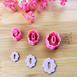 Puede incluir: Cortadores de galletas en forma de flor, rosas y lavanda, de varios tamaños. Los cortadores están dispuestos sobre una superficie de madera, con flores rosas en el fondo. Cada cortador presenta un cero en el centro.