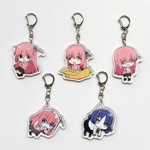 Puede incluir: Cinco llaveros de estilo anime con anillos de metal plateado. Cada llavero presenta un diseño de personaje diferente con cabello rosa o azul, vistiendo uniformes escolares o atuendos casuales. Algunos llaveros muestran personajes dentro de una caja.