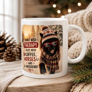 Op de afbeelding: Een witte keramische mok met een afbeelding van een paard met een wintermuts met patroon, een bijpassende sjaal en een rode zonnebril. De tekst luidt: I DON'T NEED THERAPY I JUST NEED COFFEE, HORSES AND A WEEKEND! Achtergrond met een bos in de sneeuw.