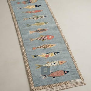 Op de afbeelding: Een rechthoekige lichtblauwe loper met een herhalend patroon van tien gestileerde vissen in verschillende kleuren en texturen. Het kleed heeft een crèmekleurige rand met een geometrisch patroon en korte franjes aan beide uiteinden.
