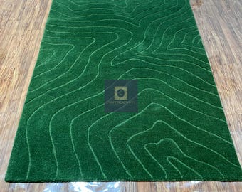 May include: A rectangular green rug with a topographic design. The rug features a series of curved lines that create a textured, layered effect. The rug is placed on a wooden floor.