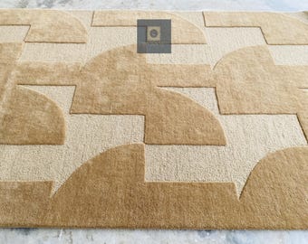 Puede incluir: Una alfombra con estampado geométrico beige y marrón claro. La alfombra presenta un patrón repetitivo de formas curvas y rectangulares en diferentes tonos de beige y marrón claro. El diseño crea un aspecto texturizado y moderno. La alfombra está hecha de un material suave.