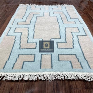 May include: A rectangular rug with a geometric pattern in light blue, beige, and dark blue. The rug has a fringe on the short ends and is placed on a wooden floor. The design features a central shape with stepped lines.