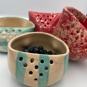 Könnte beinhalten: Eine Sammlung handgefertigter Keramik-Beeren-Schalen in verschiedenen Farben. Eine Schale ist mit Blaubeeren gefüllt. Die Schalen haben eine beige Basis mit türkisfarbenen Streifen und Löchern. Andere Schalen sind rot mit weißen Sprenkeln und Löchern sowie türkis mit weißen Sprenkeln und Löchern.