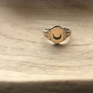 Peut inclure: Une bague en argent avec un motif de croissant de lune sur le dessus. La bague est posée sur une surface en bois brun clair.