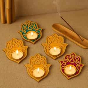 Puede incluir: Cinco candelabros en forma de mano de Hamsa en oro, turquesa y rojo. Cada soporte tiene una vela de t&eacute; encendida. Un porta incienso de madera y tallos de bamb&uacute; est&aacute;n en el fondo. Los candelabros est&aacute;n sobre una superficie de color neutro.