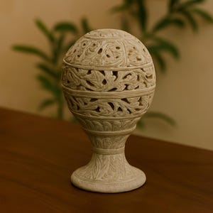 Peut inclure: Objet décoratif en pierre beige sculptée, avec un sommet sphérique et une base en piédestal. La sphère présente des découpes florales complexes, et la base a un motif de feuilles détaillé. L'objet est posé sur une surface en bois, avec une plante verte floue en arrière-plan. Hauteur: environ 20 cm.