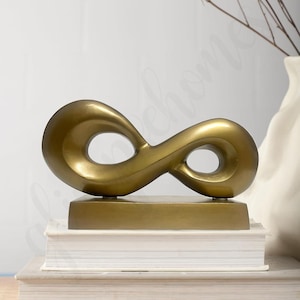 Puede incluir: Escultura dorada con forma de símbolo de infinito sobre una base rectangular, apoyada en una pila de libros. La escultura tiene una superficie lisa y reflectante, un elemento decorativo.