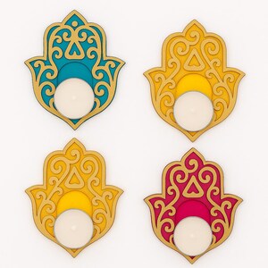 Puede incluir: Cuatro candelabros en forma de mano de Hamsa en verde azulado, amarillo y rosa con detalles dorados. Cada soporte tiene un espacio circular para una vela de t&eacute;. El dise&ntilde;o presenta intrincados patrones recortados.