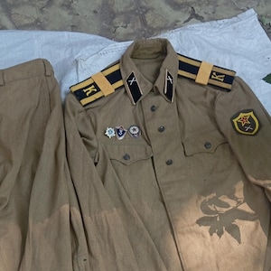 Puede incluir: Un conjunto de uniforme de estilo militar color canela que consta de pantalones largos y una chaqueta de manga larga con botones. La chaqueta presenta hombreras negras y amarillas con la letra K, cuello con pestañas negras y varios pines.