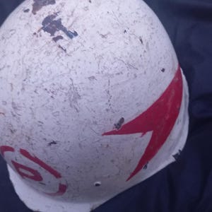 Puede incluir: Un casco blanco con diseños de flechas y círculos rojos. El casco muestra signos de desgaste, con pintura descascarillada y decoloración. El casco es probablemente para deportes o recreación.