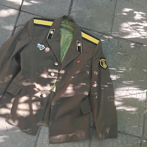 Puede incluir: Una chaqueta de estilo militar verde oliva con hombreras amarillas, solapas negras con emblemas plateados y varios pines metálicos pequeños en el pecho. La chaqueta incluye un parche en la manga izquierda y un pequeño emblema rojo en la solapa.