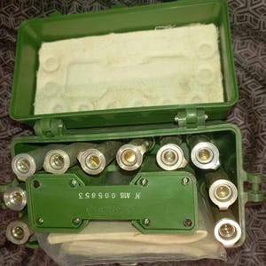 Puede incluir: Una caja de almacenamiento de plástico verde que contiene múltiples componentes cilíndricos metálicos con aberturas circulares. La caja cuenta con una tapa abatible con un inserto de espuma blanca y un panel central verde con el texto N A16 005853.