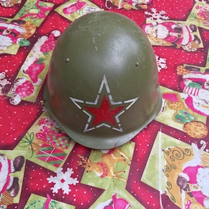 May include: An olive green military-style helmet featuring a prominent red star with a white outline centered on the front. The helmet has a smooth, rounded dome shape with small metal rivets visible near the top and shows signs of surface wear.