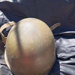 Puede incluir: Un casco militar verde oliva con una superficie desgastada y ligeramente rayada. El casco tiene una correa de color beige y está sobre una superficie oscura y texturizada. La forma del casco es redondeada.