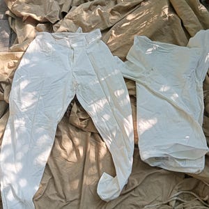 May include: A pair of off-white trousers featuring a button closure and a matching off-white long-sleeved shirt with a collar and button-down front. Both garments are made of a solid, light-colored fabric and are displayed together as a set.