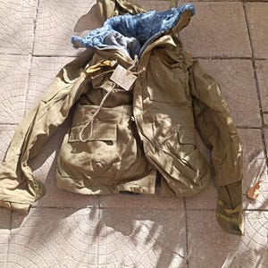 Puede incluir: Una chaqueta con capucha de color verde oliva con forro de piel sintética azul, mangas largas y varios bolsillos delanteros. Una pequeña etiqueta de papel blanco está unida al cuello con un cordel. La chaqueta tiene cuello y cremallera.
