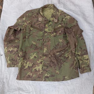 May include: A long-sleeved military-style jacket featuring a camouflage pattern in shades of olive green, brown, and tan. The jacket includes a collared neckline, two large chest pockets, and two embroidered star insignias on the collar points.