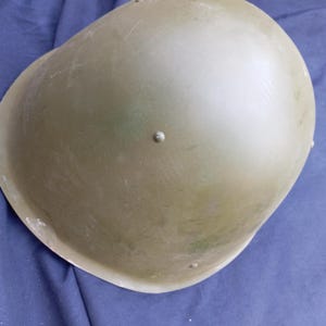 May include: An olive drab military helmet with a rounded top and a slightly textured surface. The helmet has a few small rivets and a slightly worn appearance, suggesting it may be vintage or used. The helmet is likely made of metal.