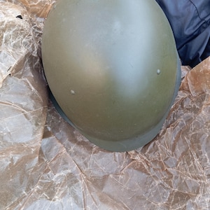 May include: An olive drab military helmet. The helmet is a rounded shape with a brim and two small rivets. It rests on crumpled brown paper.