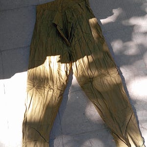May include: Olive green trousers featuring a high-waisted design with a front zip fastening and vertical stitched pleats running down the length of each leg. The trousers have a slightly creased texture and include adjustable straps at the ankles.