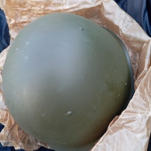 May include: An olive drab military helmet rests inside crumpled brown packing paper. The helmet has a rounded shape with a slight brim and several small rivets. The background is a dark blue fabric.