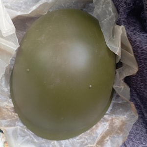 May include: An olive green military-style helmet with a smooth, rounded dome shape. The helmet features two small, raised circular rivets on the exterior surface and is partially wrapped in translucent, crinkled protective plastic packaging.